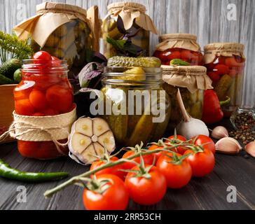 Pomodori ciliegini in scatola e cetriolini in barattoli, verdure fresche, spezie ed erbe per marinare su uno sfondo di legno. Foto Stock