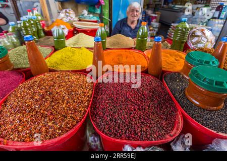 Un commerciante nella sua stalla di spezie e frutta secca al Dezerter Bazaar di Tbilisi, Georgia. In primo piano ci sono grandi ciotole di barbacche essiccate (Berberis vulgaris), utilizzate nella cucina georgiana per condire piatti di carne e salse, nonché altri frutti secchi; dietro il commerciante ci sono polveri di spezie colorate e bottiglie di essenze. Il Dezerter Bazaar è sotto minaccia di demolizione dal 2024. Mikheil Tsinamdzghvrishvili Street, Chughureti, Didube, Tbilisi, 0102, Georgia Foto Stock