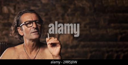 Un uomo più anziano, di circa 60 anni, di nazionalità serba, con capelli lunghi e occhiali, fuma volutamente una sigaretta su uno sfondo scuro. Artista, cre Foto Stock