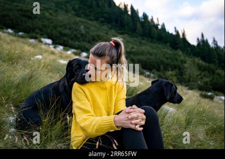 Giovane proprietaria di due cani labrador retriever neri di razza beautfiul seduti in erba verde sulla collina con uno dei suoi cani appoggiati alla testa Foto Stock