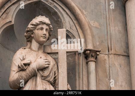 antica statua religiosa in marmo di donna che porta croce cristiana in un cimitero all'aperto Foto Stock