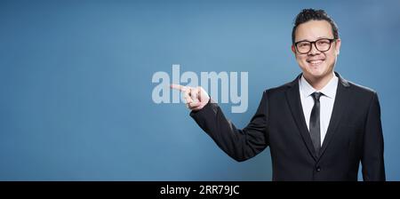 Ritratto di imprenditore asiatici sorriso e punti con il dito a sinistra della videocamera su vintage sfondo blu . Foto Stock