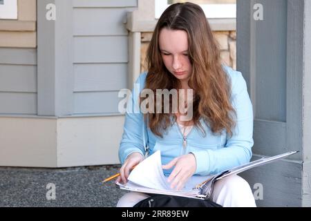 giovane ragazza adolescente siede su portico in mani con una cartella, rivista e matita, fare i compiti a casa, risolvere i problemi, attendere alla porta, una bella donna, giovani jeans bianchi, una valigetta, pensare, lavoro, studio Foto Stock