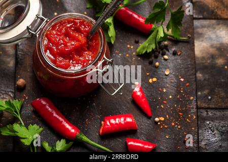 Salsa piccante fatta in casa al peperoncino in un piccolo vaso di vetro su un tavolo da cucina in legno scuro, con peperoni rossi caldi e prezzemolo Foto Stock