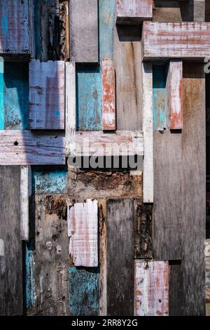 Struttura di sfondo con tavole di legno intemprate con resti grintosi di vernice blu, rossa e marrone che formano il lato di un edificio esterno wal Foto Stock