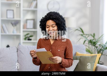 Una giovane donna afroamericana sorridente seduta a casa e che apre una lettera dalla busta, ha ricevuto notizie, un messaggio. Foto Stock