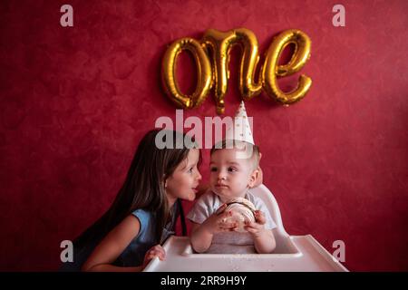 La sorella maggiore si congratula con il fratello minore per il suo primo compleanno. La ragazza bacia gli abbracci da bambino. I bambini si divertono su sfondo rosso con la lamina B. Foto Stock