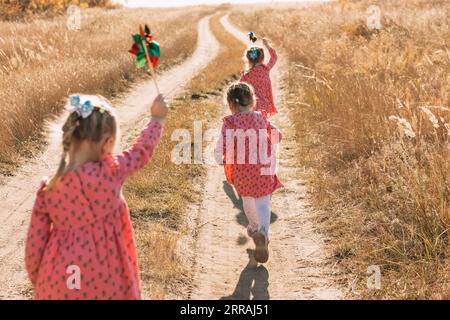 Giocare nel vento: Le ragazze triplet corrono verso avventure con i mulini a vento in una calda giornata autunnale, utilizzando l'energia eolica Foto Stock