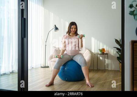 La radiosa mamma asiatica abbraccia il benessere, scolpendo la forza nel suo santuario di casa. I manubri diventano strumenti di empowerment nel suo grazioso viaggio. Foto Stock