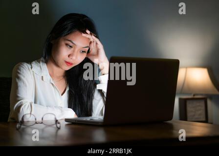 Una donna affaticata e sovraccarica tiene la testa in mano mentre lavora e studia su un personal computer. Foto Stock
