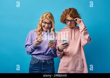 allegra ragazza bionda con occhiali da vista che guarda lo smartphone vicino a un'amica rossa su blu Foto Stock