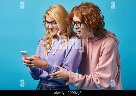 gioiosa ragazza bionda con occhiali da vista e felpa con cappuccio con smartphone vicino a un amico rosso su blu Foto Stock