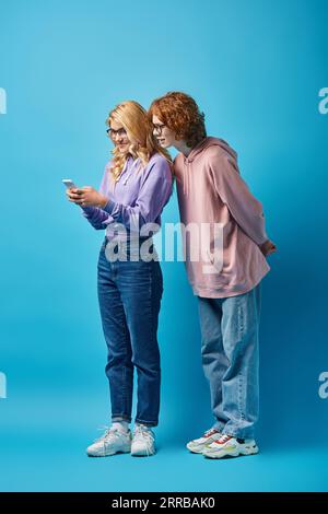 ragazza adolescente sorridente con occhiali da vista e abiti eleganti utilizzando lo smartphone vicino a un amico rosso su blu Foto Stock