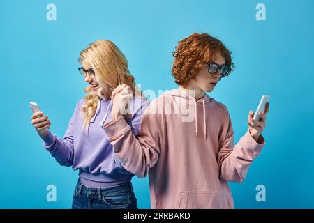 gli amici adolescenti indossano occhiali da vista e felpe con cappuccio eleganti utilizzando i telefoni cellulari e tenendo le mani sul blu Foto Stock