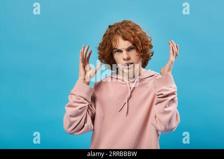 ragazzo adolescente irritato con i capelli rossi ondulati che gestiscono e guardano la fotocamera in blu, di cattivo umore Foto Stock