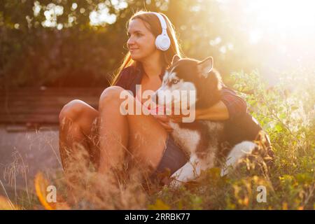 La bella giovane donna con le cuffie gioca, abbraccia con il suo cane all'aperto al tramonto Foto Stock