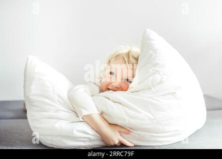 l'incantevole bambina bionda con un vestito bianco gioca con un cuscino sul letto a casa. piccola ragazza affascinante giace su un cuscino bianco. Tessuti accoglienti per Foto Stock