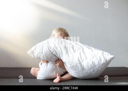 bambino piccolo nascosto dietro un cuscino bianco. Gambe e mani sporgono da dietro il cuscino dei raggi solari. affascinante bambina bionda con un vestito bianco Foto Stock