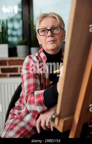 Corsi d'arte per anziani. Donna matura pensiva seduta di fronte al cavalletto guardando da parte, ascoltando l'insegnante durante la lezione di disegno, imparando a disegnare in piccoli gruppi. Persone anziane e creatività Foto Stock