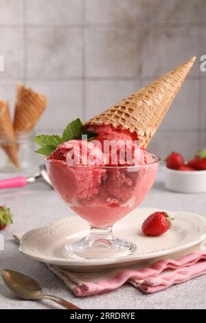 Deliziose palline di gelato alla fragola con cono di menta e cialda in un recipiente da dessert in vetro servito su un tavolo grigio Foto Stock