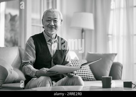 vecchio asiatico seduto sul divano a casa a leggere un libro che guarda la fotocamera sorridente, bianco e nero Foto Stock
