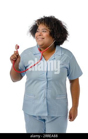 giovane medico afro latina in uniforme blu, in piedi con il suo stetoscopio rosso, in mano guardando lateralmente, immagine verticale su sfondo bianco, st Foto Stock