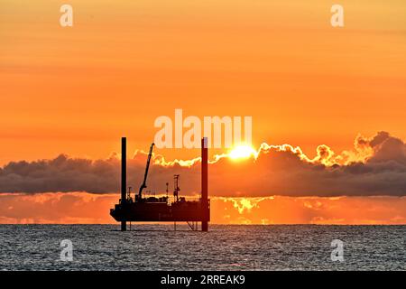 Carro di prova di perforazione ARAN250 jackup con orizzonte dorato cielo arancio nuvole e sole che sbircia sopra le nuvole nel Mare del Nord all'alba con cielo caldo mare e S. Foto Stock