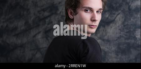 uomo dai capelli castani chiari, con una camicia nera, si erge con cura davanti a una lavagna grunge. Il suo sguardo introspettivo offre un'ampia gamma di contenuti COPYSPACE Foto Stock