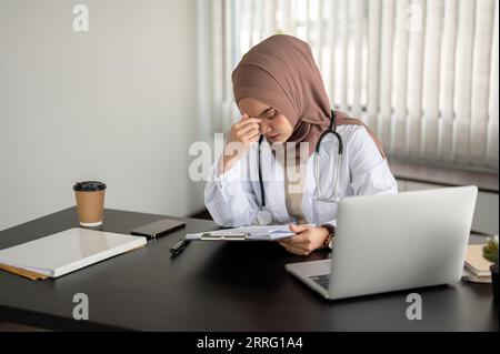 Una premurosa e stressata dottoressa musulmana asiatica in un abito bianco e hijab si sente stanca mentre lavora alla sua scrivania nel suo ufficio in un ospedale. Foto Stock