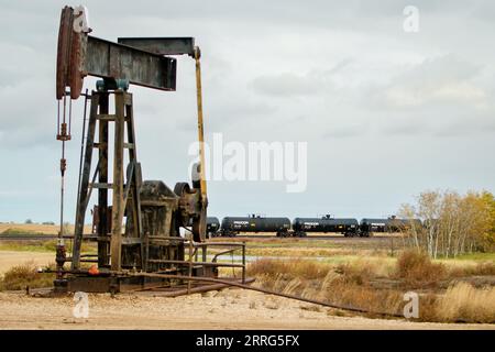 Un martinetto pompa pompa l'olio proveniente da un pozzetto di olio di scisto all'esterno di Williston, una sezione del campo dell'olio di Bakken. Sullo sfondo, un treno merci con carri cisterna pieni di petrolio. Foto Stock