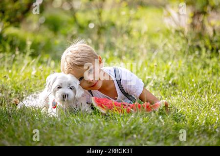 Fantastico bambino biondo, ragazzo con cane, anguria in giardino, estate Foto Stock