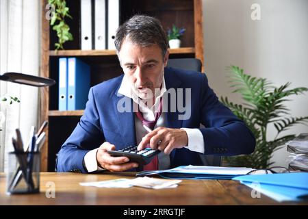 Dettaglio di un uomo d'affari preoccupato e sorpreso che controlla i conti della sua azienda su una calcolatrice sul tavolo del suo ufficio Foto Stock