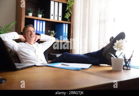 Uomo seduto felice nel suo ufficio con i piedi sul tavolo di legno con documenti e scaffali con armadietti dietro Foto Stock