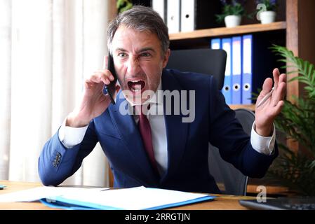 Uomo d'affari che parlava al telefono arrabbiato e urlava nel suo ufficio seduto con documenti sul tavolo in ufficio con mobili di legno Foto Stock