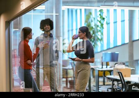 Tra le moderne pareti di vetro di un ufficio startup, un gruppo eterogeneo di colleghi, tra cui un uomo afroamericano, si impegnano in un caffè alimentato Foto Stock