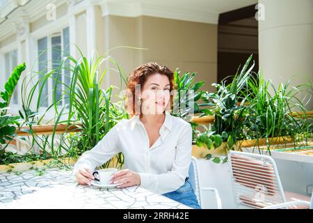 Bella donna alla moda dai capelli rossi beve caffè in un caffè. Foto Stock