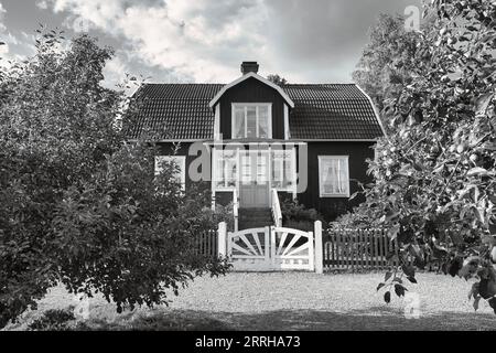 una tipica casa svedese rossa e bianca in piccolo bianco e nero. Cancello bianco del giardino, recinzione marrone. prato verde in giardino. Alberi in primo piano Foto Stock