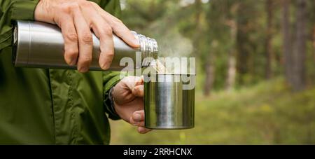 l'uomo versa il tè caldo dal thermos in una tazza nella foresta. turismo naturalistico e campeggio. striscione con spazio per la copia Foto Stock