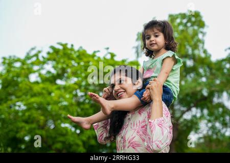 Giovane madre indiana felice che porta la figlia sulle spalle al parco estivo. Concetto di famiglia e maternità. Foto Stock