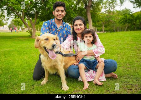 Felice giovane famiglia indiana che si diverte insieme al parco estivo. Madre, padre e figlia con cane labrador in giardino. Foto Stock