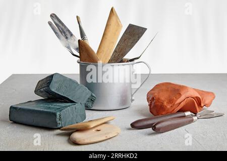 Strumenti per modellare e modellare creativi in una tazza di alluminio e pezzi di argilla e plasticina su un tavolo di cemento bianco in un laboratorio di scultori Foto Stock