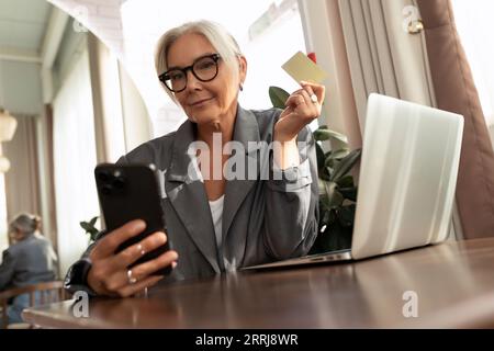 una donna di mezza età ben curata e di successo con gli occhiali paga un ordine utilizzando uno smartphone e una carta di credito in un bar Foto Stock