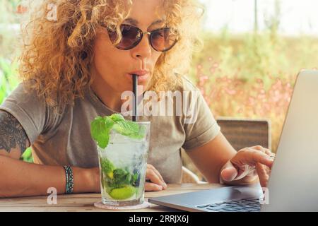 Lavoro estivo e online nuovo stile di vita moderno per il business con una donna che beve cocktail con paglia e lavora scrivendo su un computer portatile al bar sulla spiaggia del caffè durante le vacanze estive. Ufficio alternativo Foto Stock