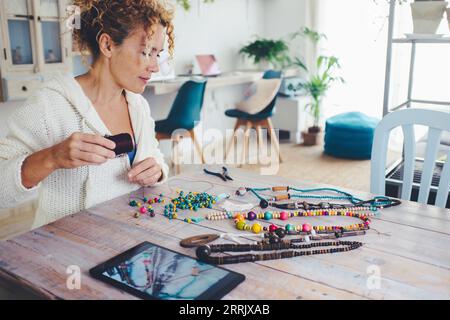 Persone che praticano attività ricreative al coperto. Una donna adulta che fa collana di perline sul tavolo in salotto con un tablet acceso. Un'alternativa moderna, il nuovo business lavora online vendendo sul Web Foto Stock