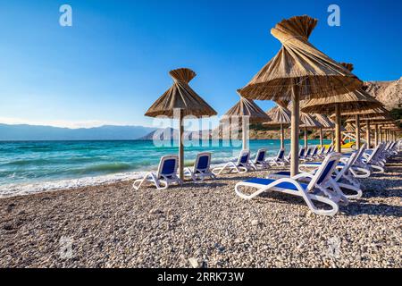 Croazia, baia del Quarnero, Primorje Gorski Kotar County, isola di Krk, lettini e ombrelloni in paglia sulla spiaggia di ciottoli di Baska Foto Stock