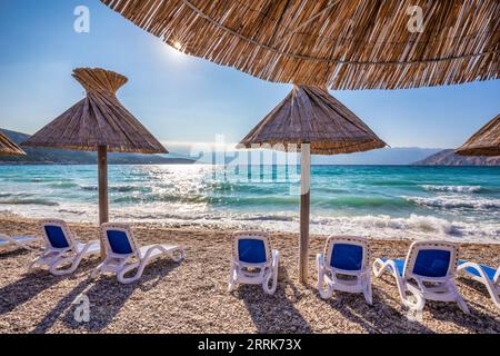 Croazia, baia del Quarnero, Primorje Gorski Kotar County, isola di Krk, lettini e ombrelloni in paglia sulla spiaggia di ciottoli di Baska Foto Stock