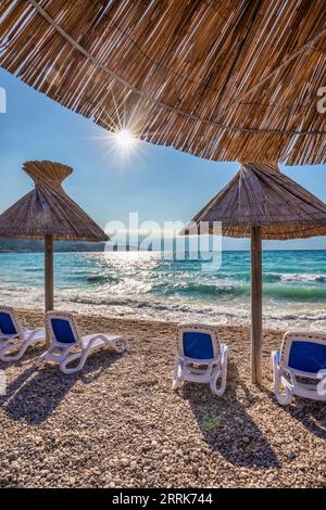 Croazia, baia del Quarnero, Primorje Gorski Kotar County, isola di Krk, lettini e ombrelloni in paglia sulla spiaggia di ciottoli di Baska Foto Stock