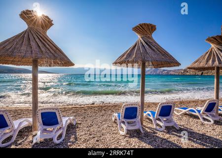 Croazia, baia del Quarnero, Primorje Gorski Kotar County, isola di Krk, lettini e ombrelloni in paglia sulla spiaggia di ciottoli di Baska Foto Stock