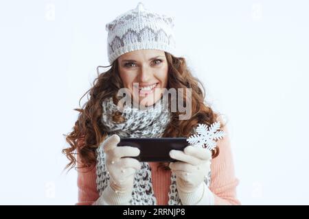 Ciao inverno. Ritratto di una donna sorridente di 40 anni con maglione, guanti, cappello e sciarpa con fiocco di neve utilizzando applicazioni per smartphone isolate Foto Stock