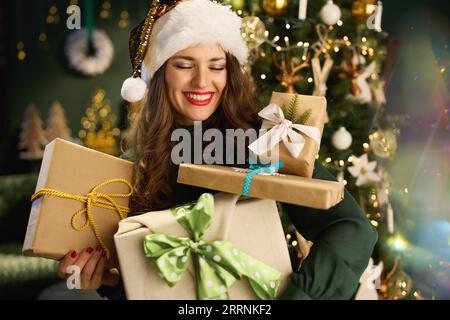 Natale. Casalinga elegante e sorridente, con cappello da Babbo Natale e scatole regalo confezionate ecologiche in abito verde vicino all'albero di Natale in una casa moderna. Foto Stock